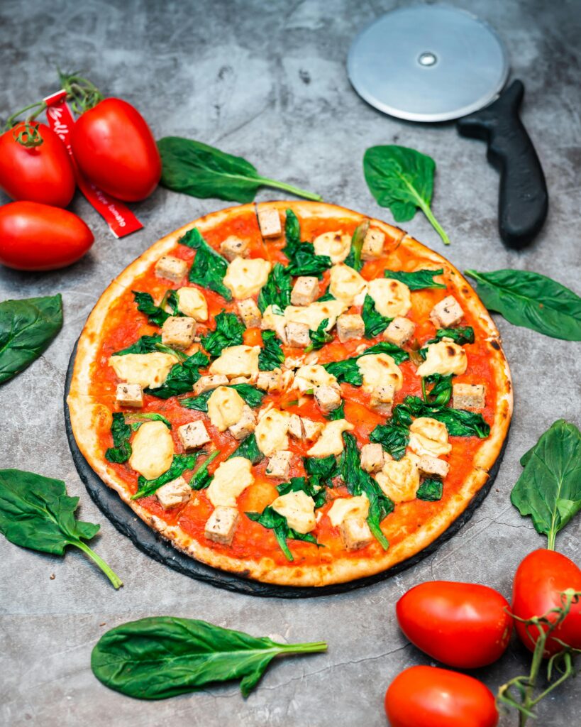 pexels-photo-10779645-10779645 Bright and fresh pizza topped with tofu, spinach, and mozzarella surrounded by tomatoes and spinach leaves.